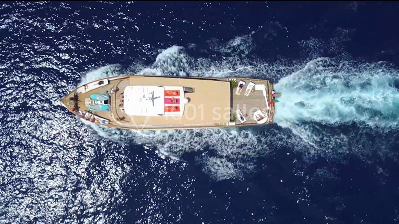 Vue aérienne d'un yacht naviguant sur une mer bleue, laissant une traînée d'écume blanche derrière lui.