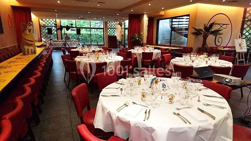 Salle de réception avec tables rondes dressées, nappes blanches, chaises rouges et décoration lumineuse.