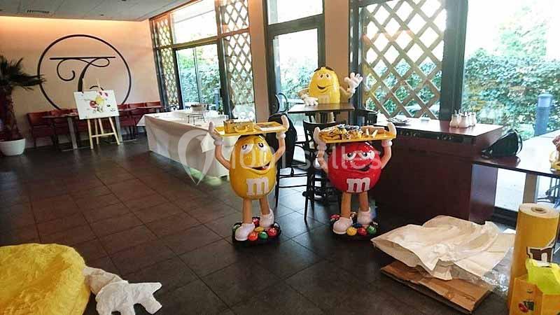Statues des personnages M&M's tenant des plateaux dans une salle lumineuse avec des tables et des décorations.
