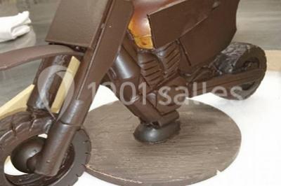 Moto miniature sculptée en chocolat, posée sur un socle rond, dans un environnement de cuisine professionnelle.