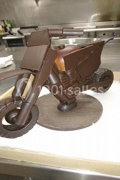 Moto miniature sculptée en chocolat, posée sur un socle rond, dans un environnement de cuisine professionnelle.