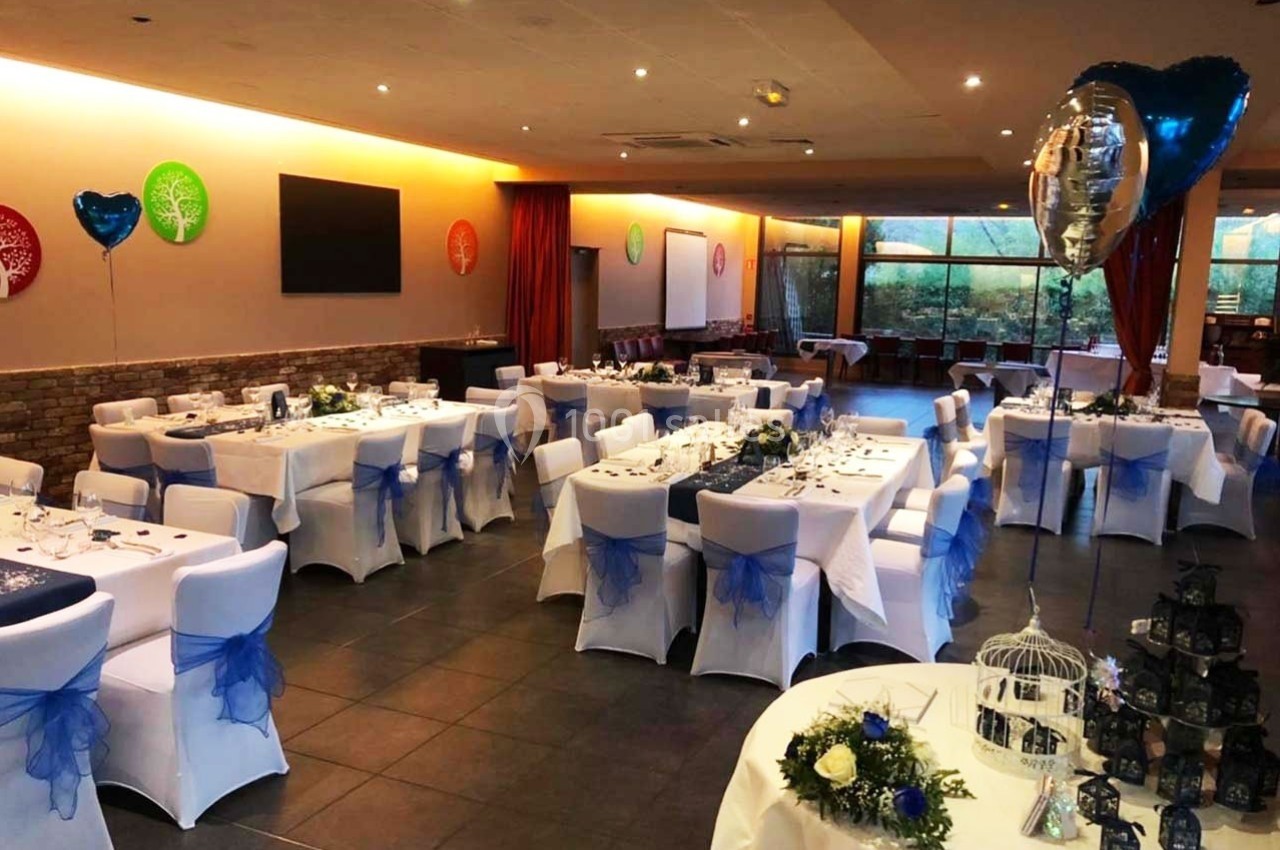 Salle de réception décorée avec des tables dressées, des chaises blanches ornées de nœuds bleus et des ballons.