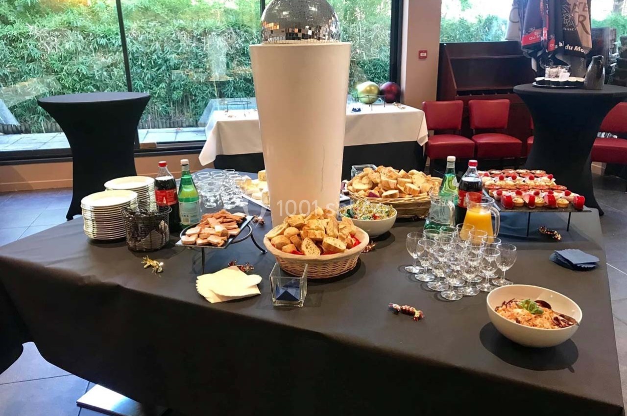 Table de buffet avec pains, boissons, verrines et salades, dans une salle avec baies vitrées et fauteuils rouges.
