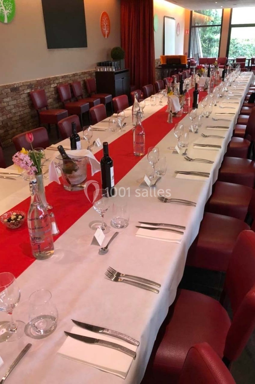 Salle de réception avec une longue table dressée, nappes blanches et rouges, couverts alignés et bouteilles de vin.