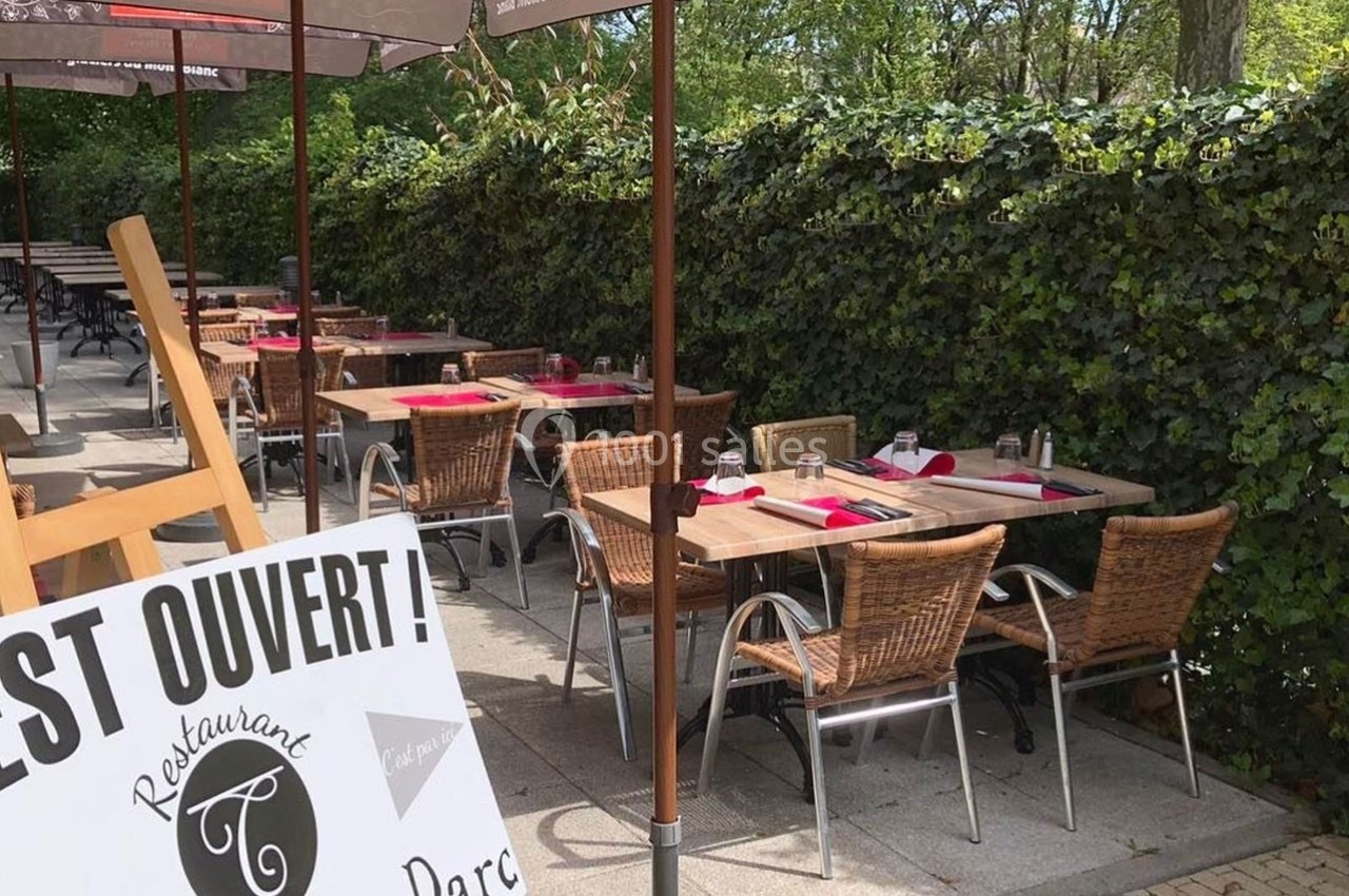 Terrasse d'un restaurant avec des tables dressées et des chaises en rotin, entourée de verdure et protégée par des parasols.