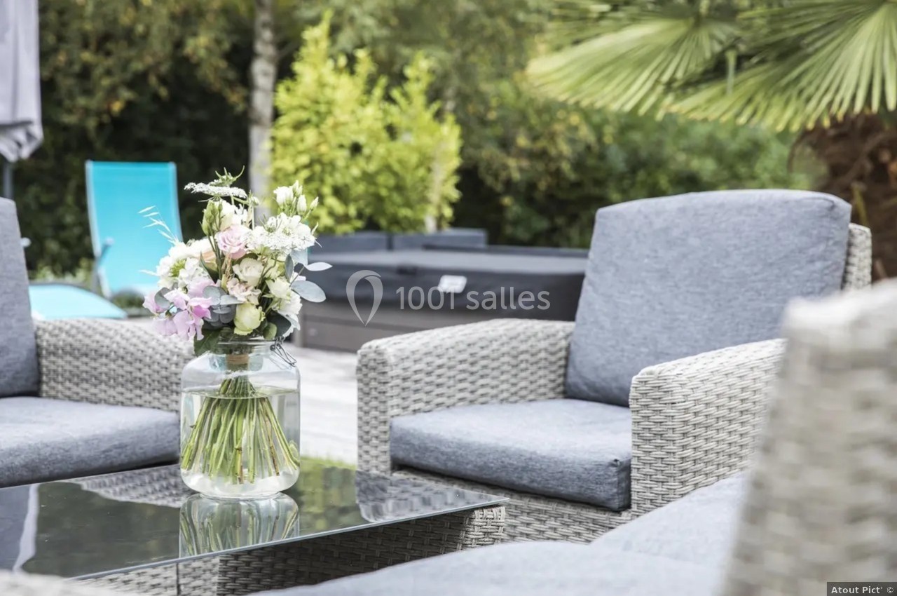 Bouquet de fleurs dans un vase en verre posé sur une table en verre, entouré de fauteuils en rotin gris dans un jardin.