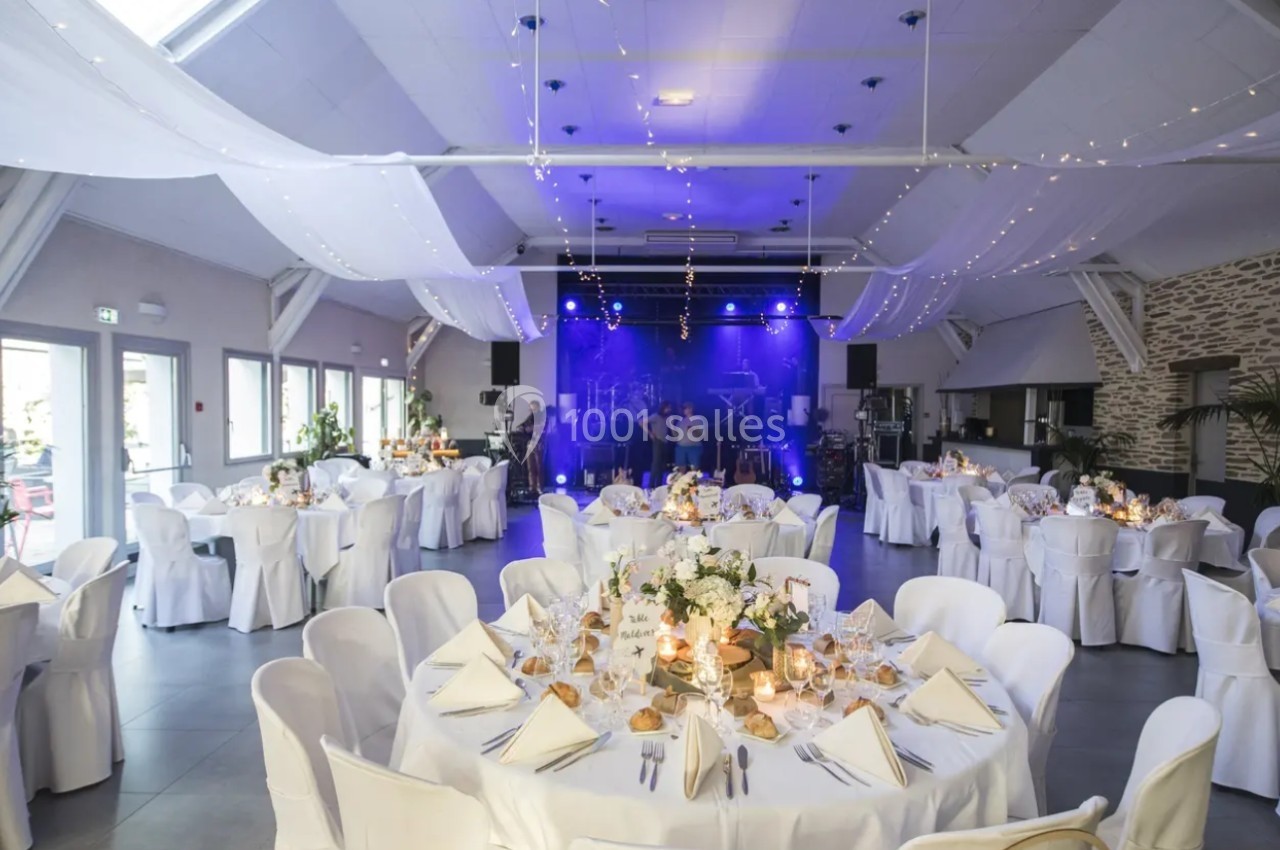 Salle de réception décorée pour un événement, avec tables dressées et scène éclairée en arrière-plan.