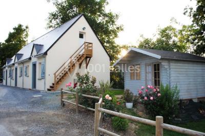 Maison blanche à étage avec escalier extérieur en bois, cabanon en bois à côté, jardin fleuri et clôture en bois.