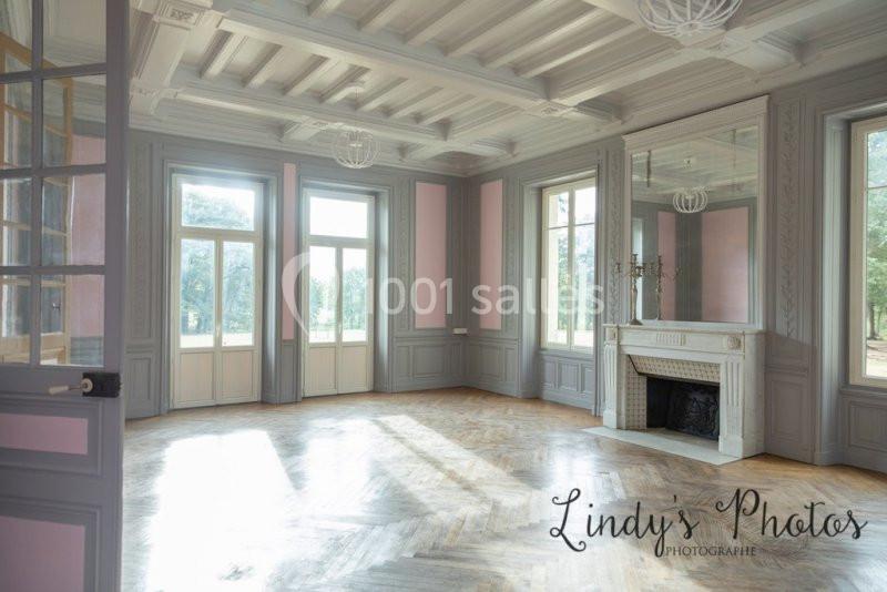 Salle lumineuse avec parquet en bois, murs gris et roses, cheminée blanche et grandes fenêtres donnant sur un jardin.