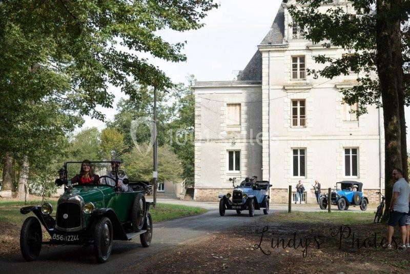 Des voitures anciennes circulent sur une allée bordée d'arbres devant un grand bâtiment en pierre.