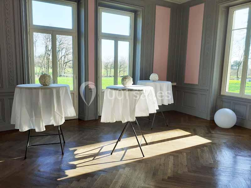 Trois tables hautes recouvertes de nappes blanches dans une pièce lumineuse avec parquet et grandes fenêtres donnant sur un…