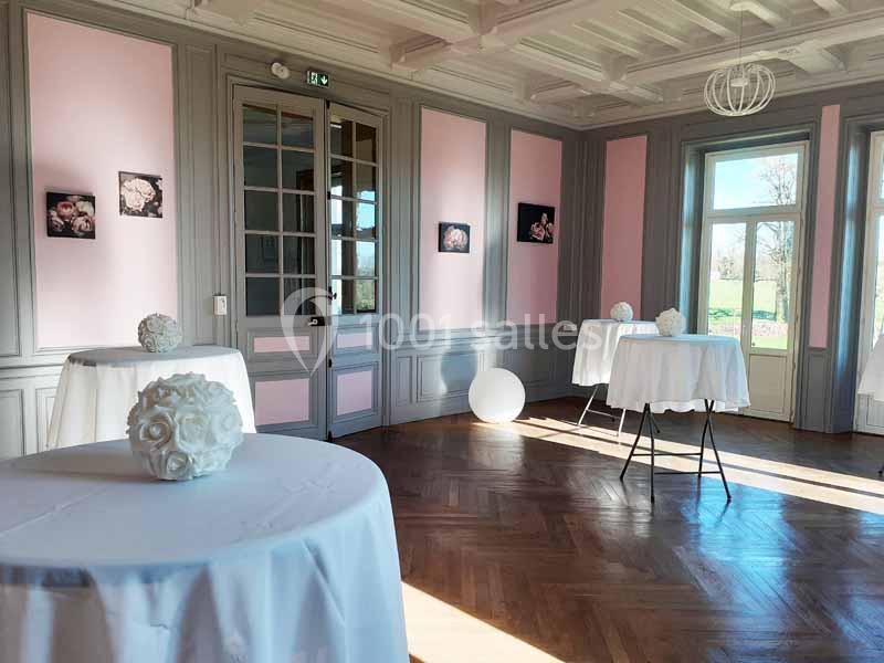 Salle lumineuse avec tables hautes décorées de bouquets blancs, murs roses et gris, et grandes fenêtres donnant sur un…