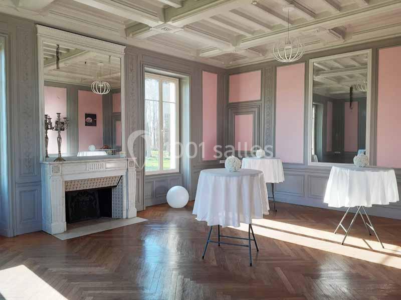 Salle lumineuse avec parquet, murs roses et gris, cheminée décorative, tables hautes rondes et grandes fenêtres.