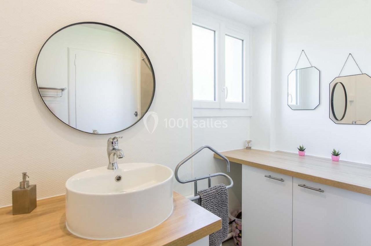 Salle de bain lumineuse avec lavabo rond, miroir mural, fenêtre, et plan de travail en bois clair.