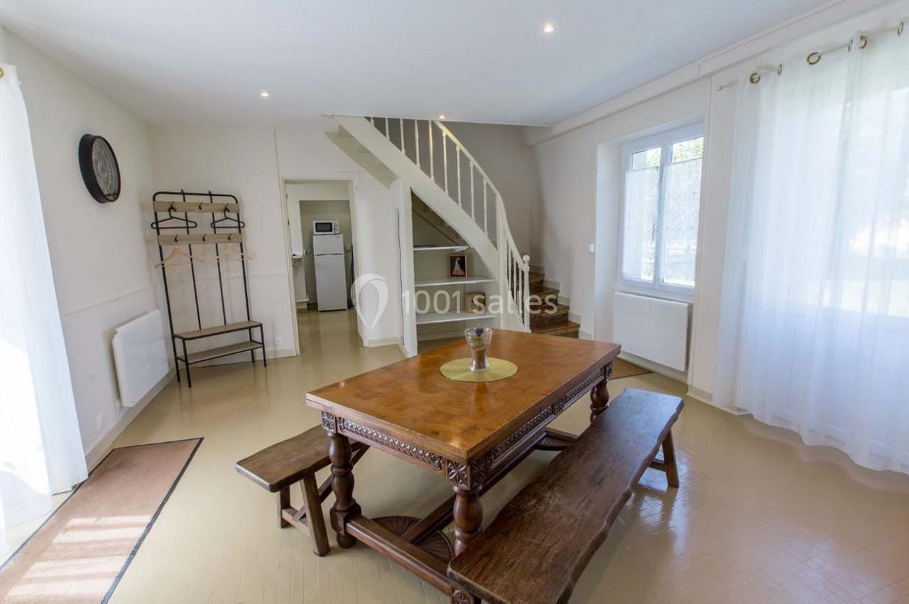 Salle à manger lumineuse avec table en bois massif, bancs assortis, escalier blanc et fenêtres avec rideaux clairs.