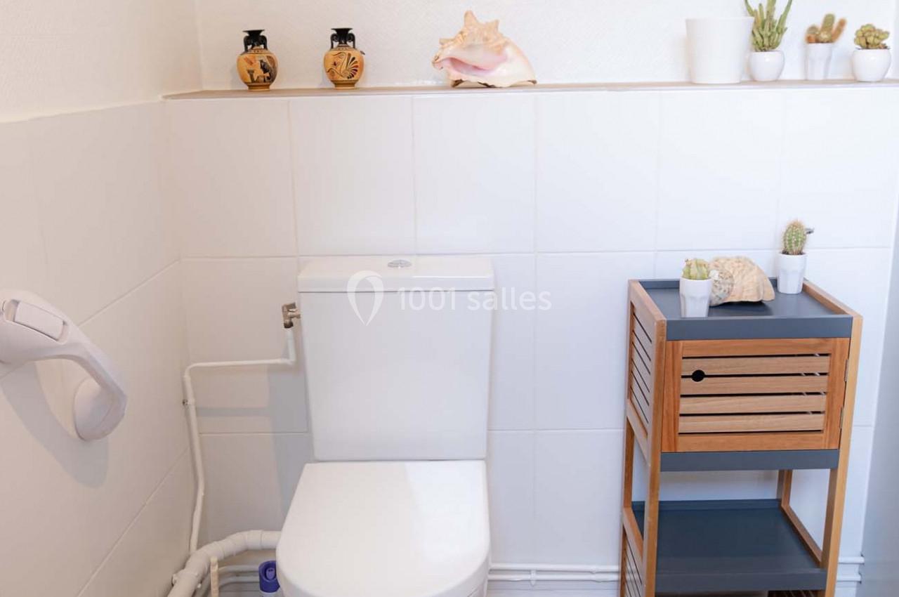 Toilettes blanches avec étagère en bois, décorée de plantes et d'objets marins, dans un espace lumineux.