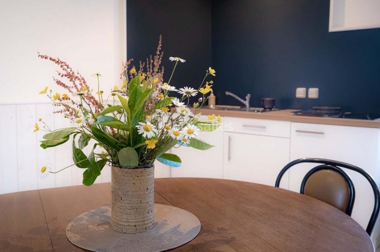 Un bouquet de fleurs sauvages dans un vase en céramique posé sur une table en bois, avec une cuisine en arrière-plan.