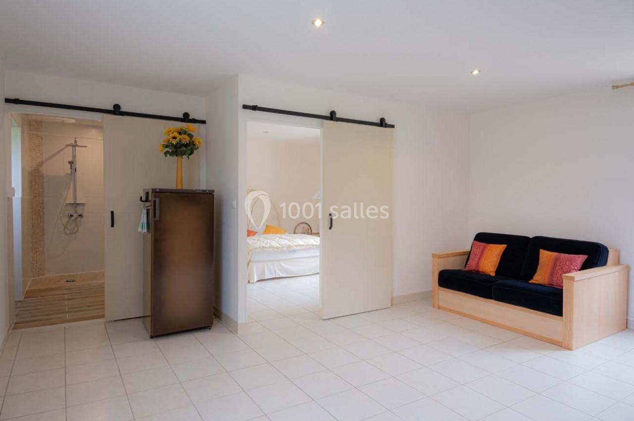 Pièce lumineuse avec canapé, lit visible derrière des portes coulissantes et salle de bain attenante.