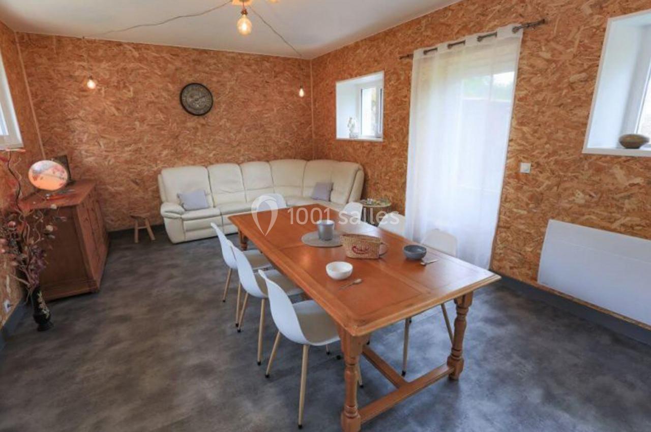 Salle à manger avec table en bois, chaises blanches, canapé beige et murs en panneaux de bois aggloméré.
