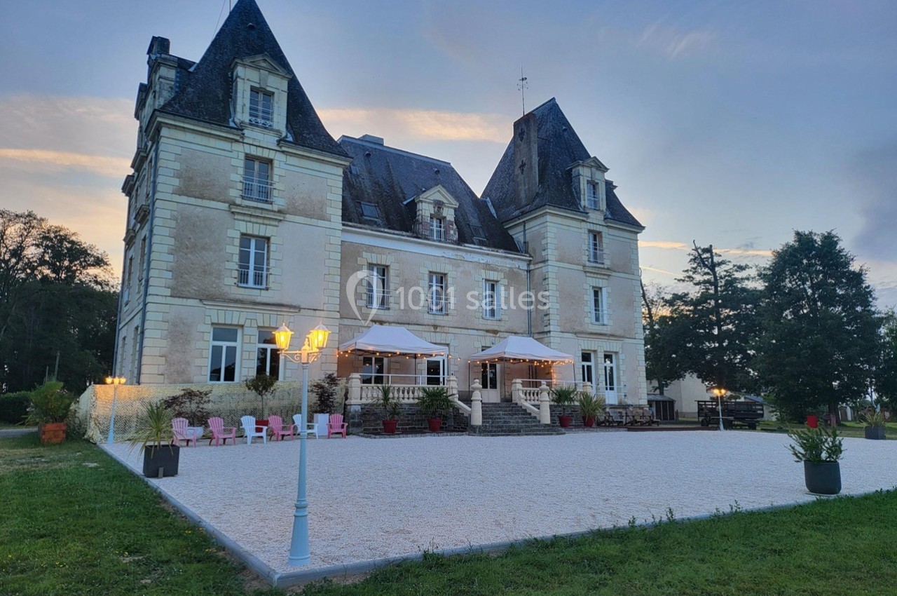 Façade d'un château en pierre avec terrasses éclairées au crépuscule, entouré de verdure et d'une cour gravillonnée.