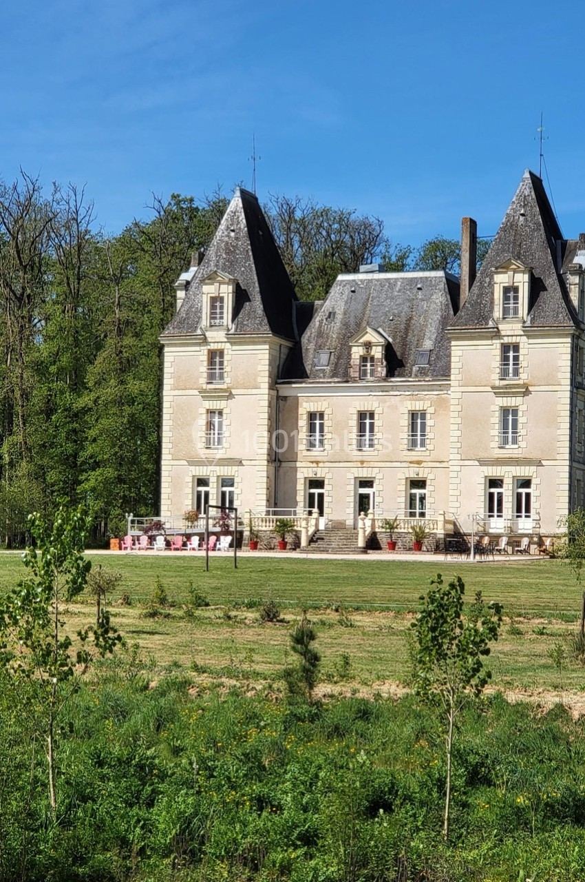 Château en pierre claire entouré de verdure, avec une terrasse aménagée et une forêt en arrière-plan.