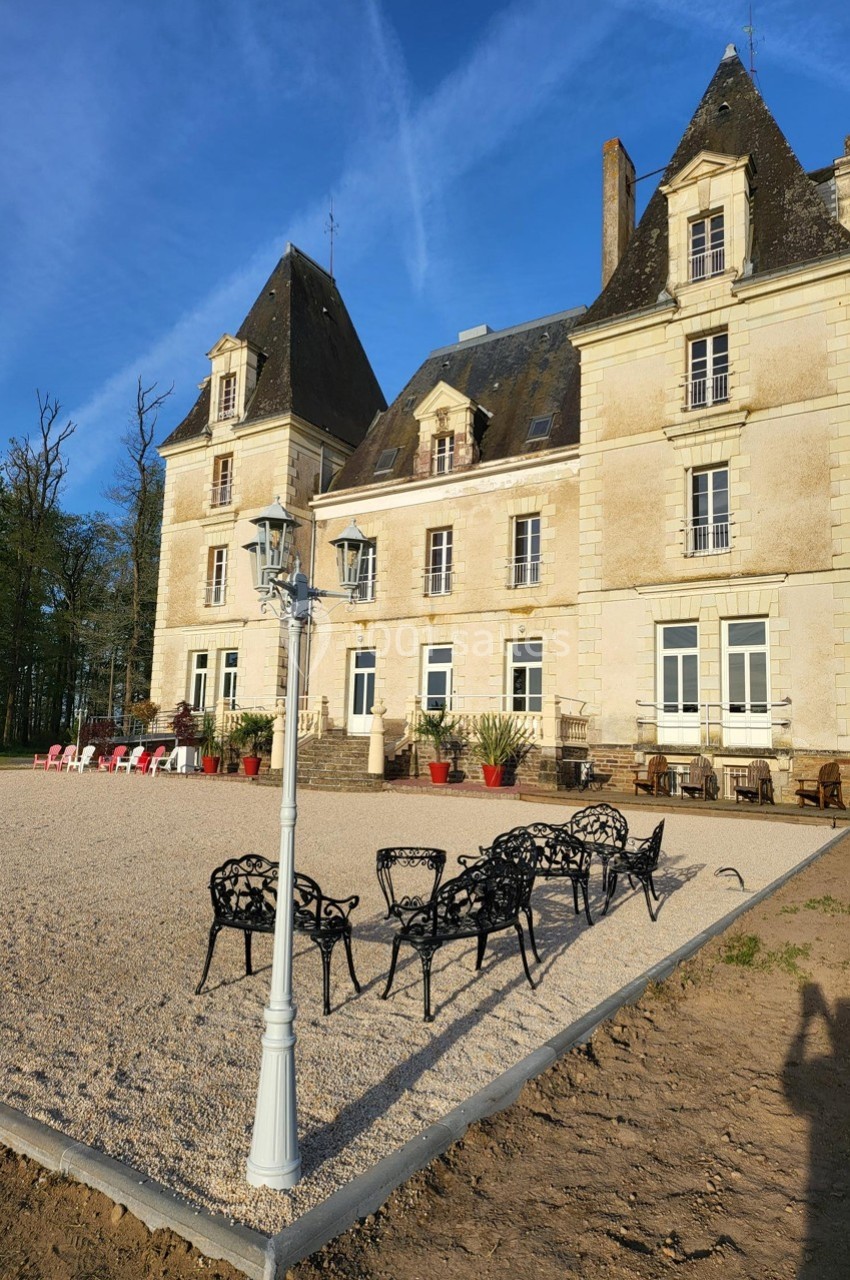 Façade d'un château en pierre avec des toits en ardoise, terrasse aménagée et mobilier extérieur sur une cour gravillonnée.