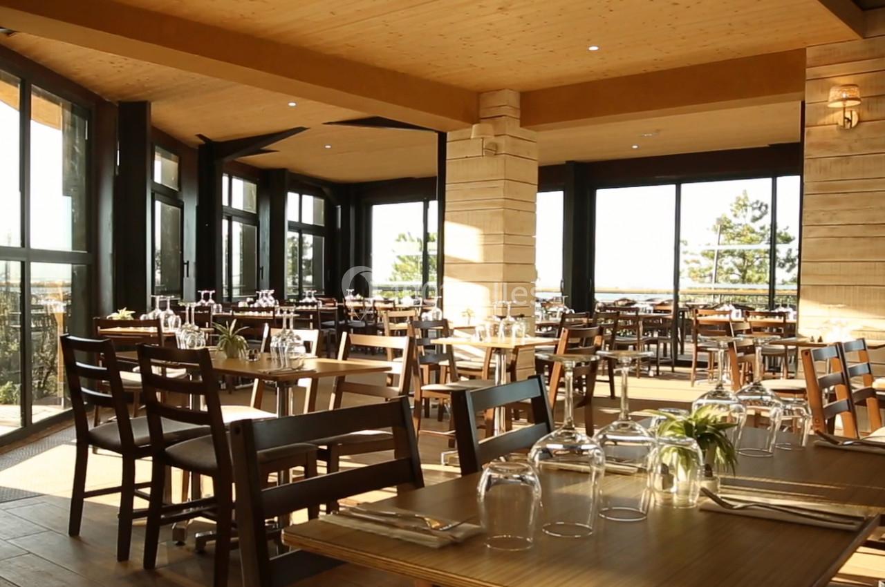 Salle de restaurant lumineuse avec de grandes baies vitrées, des tables en bois dressées et une décoration moderne.