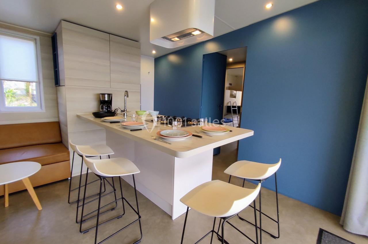 Cuisine moderne avec îlot central, tabourets hauts, mur bleu, et équipements comme cafetière et plaques de cuisson.