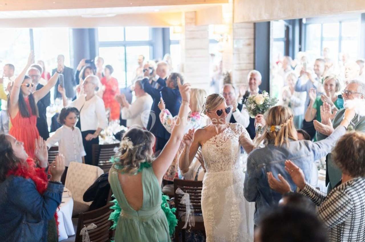 Une mariée en robe blanche entre dans une salle festive, entourée d'invités applaudissant et célébrant.