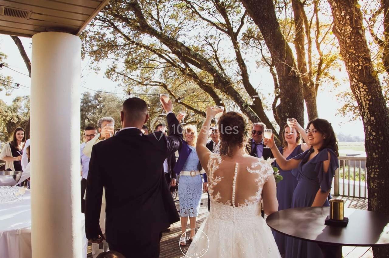 Un couple de mariés lève un verre avec des invités lors d'une réception en extérieur sous des arbres.