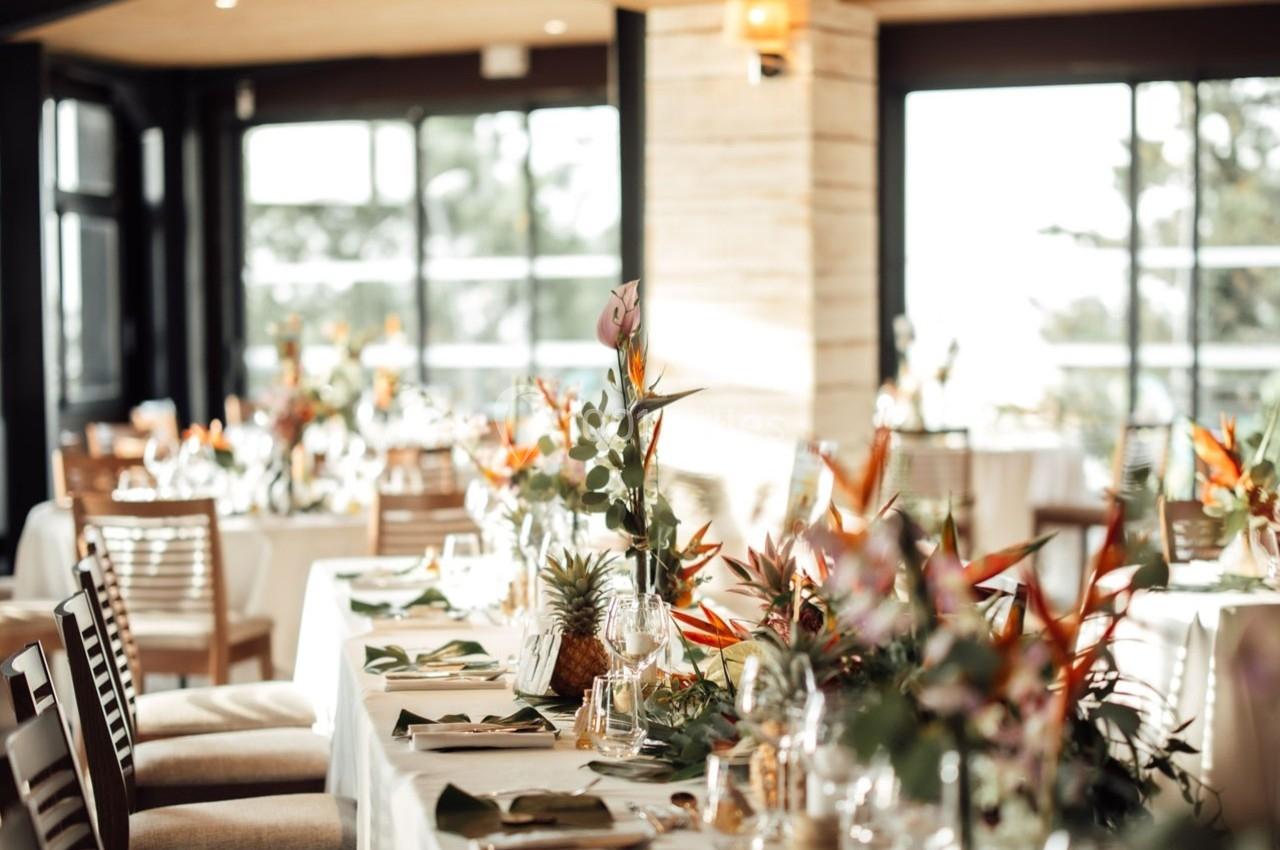 Salle lumineuse avec tables élégamment dressées, décorées de fleurs tropicales et entourées de chaises en bois.