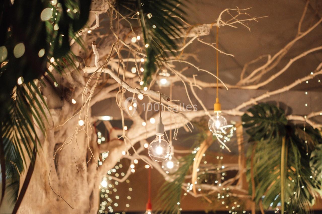 Un arbre décoratif illuminé de guirlandes lumineuses dans un espace intérieur avec des plantes et des convives en arrière…