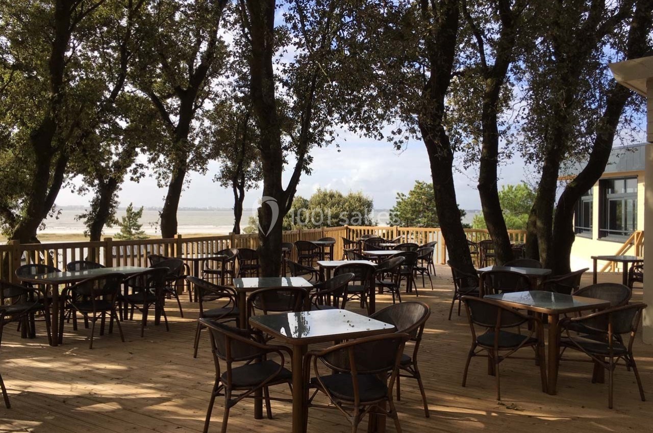 Terrasse en bois avec des tables et chaises en rotin, entourée d'arbres, offrant une vue sur un paysage naturel.