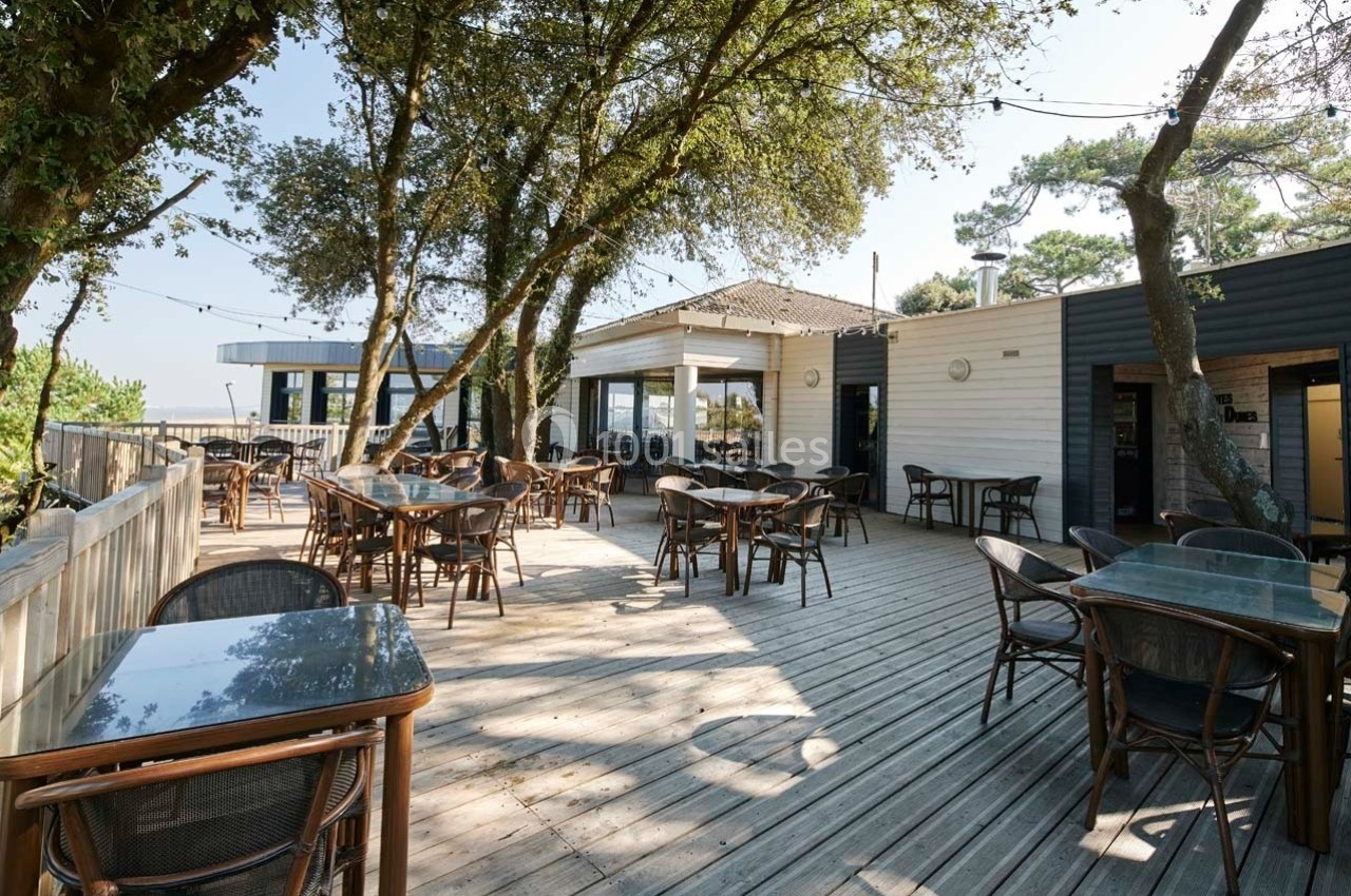 Terrasse en bois avec tables et chaises, entourée d'arbres, donnant sur un bâtiment moderne sous un ciel dégagé.