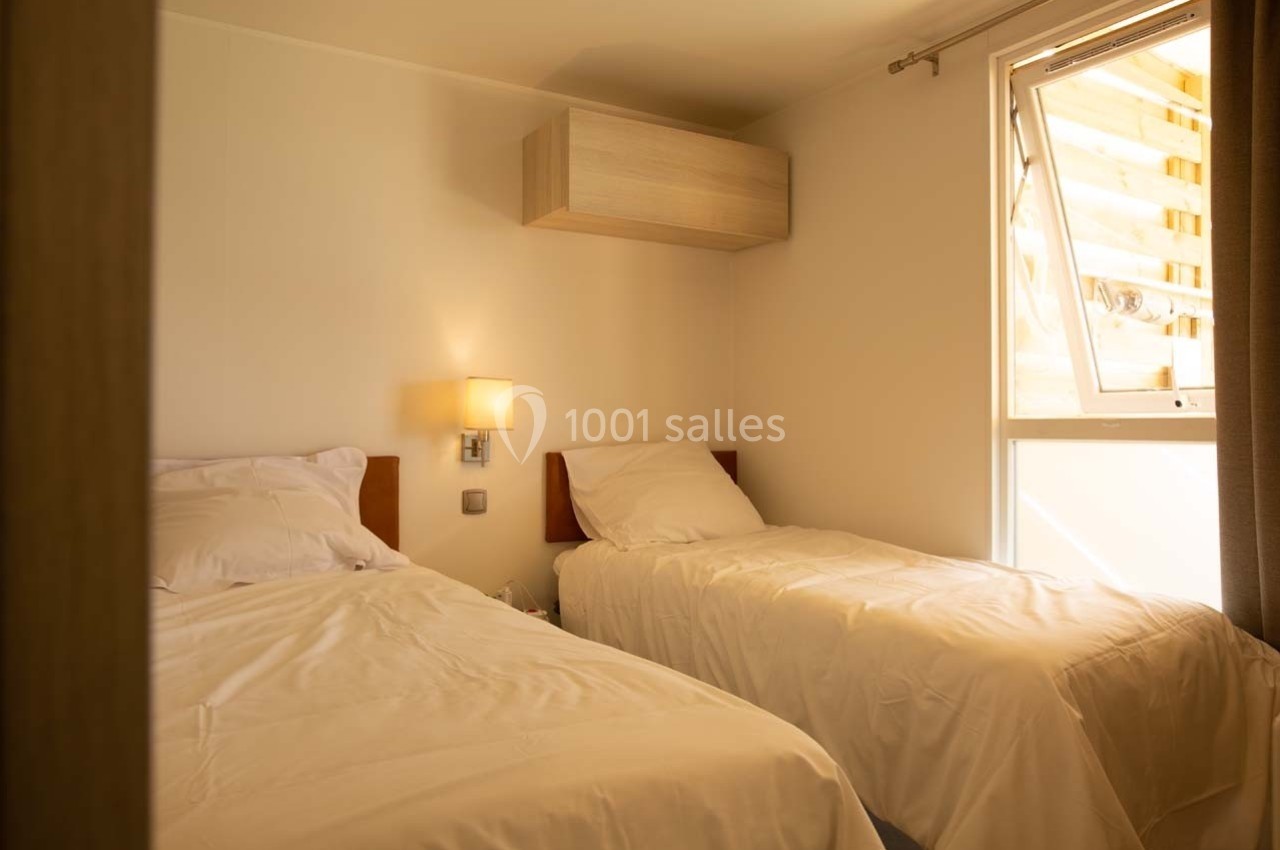 Deux lits simples avec draps blancs dans une chambre lumineuse, éclairée par une lampe murale et une fenêtre.