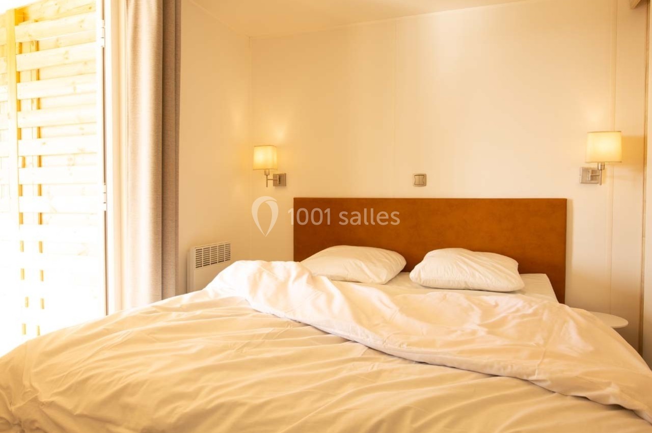 Lit double avec draps blancs défaits, encadré par deux appliques murales dans une chambre lumineuse.