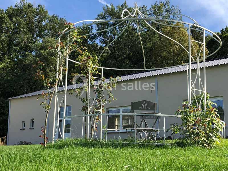 Structure métallique d'une tonnelle entourée de plantes grimpantes, devant un bâtiment beige et un jardin verdoyant.