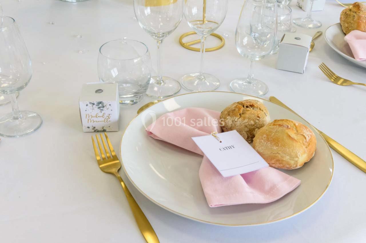 Assiette avec serviette rose pliée, deux petits pains, couverts dorés et décoration de table élégante.