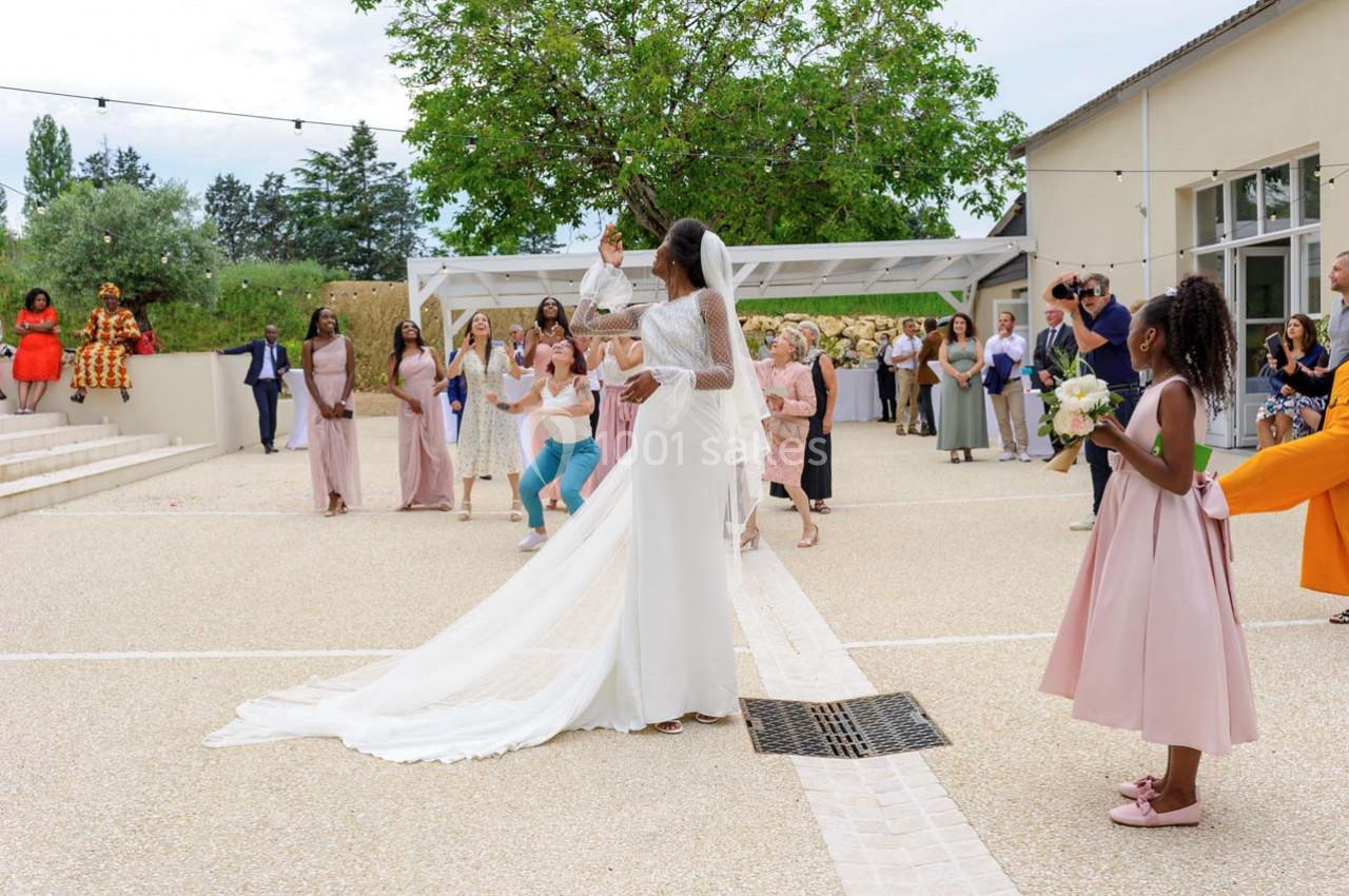 Une mariée lance son bouquet à un groupe d'invitées dans une cour extérieure lors d'une réception de mariage.