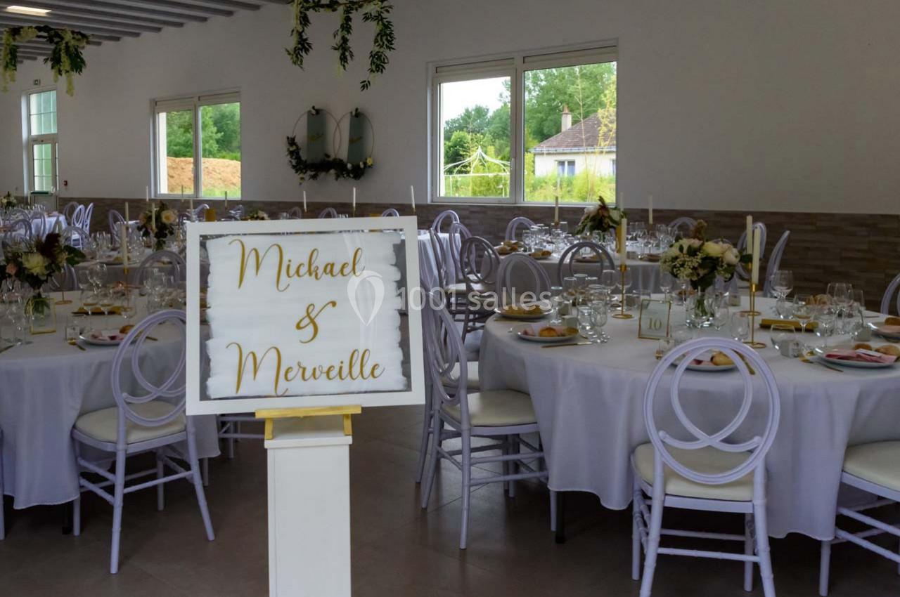 Salle de réception décorée pour un mariage, avec des tables dressées et un panneau portant des prénoms.