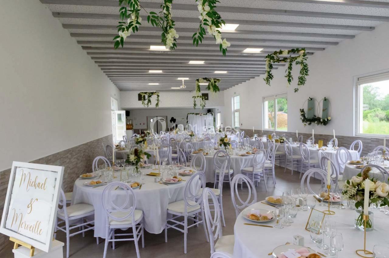 Salle de réception décorée pour un mariage, avec tables rondes, chaises blanches et ornements floraux suspendus.
