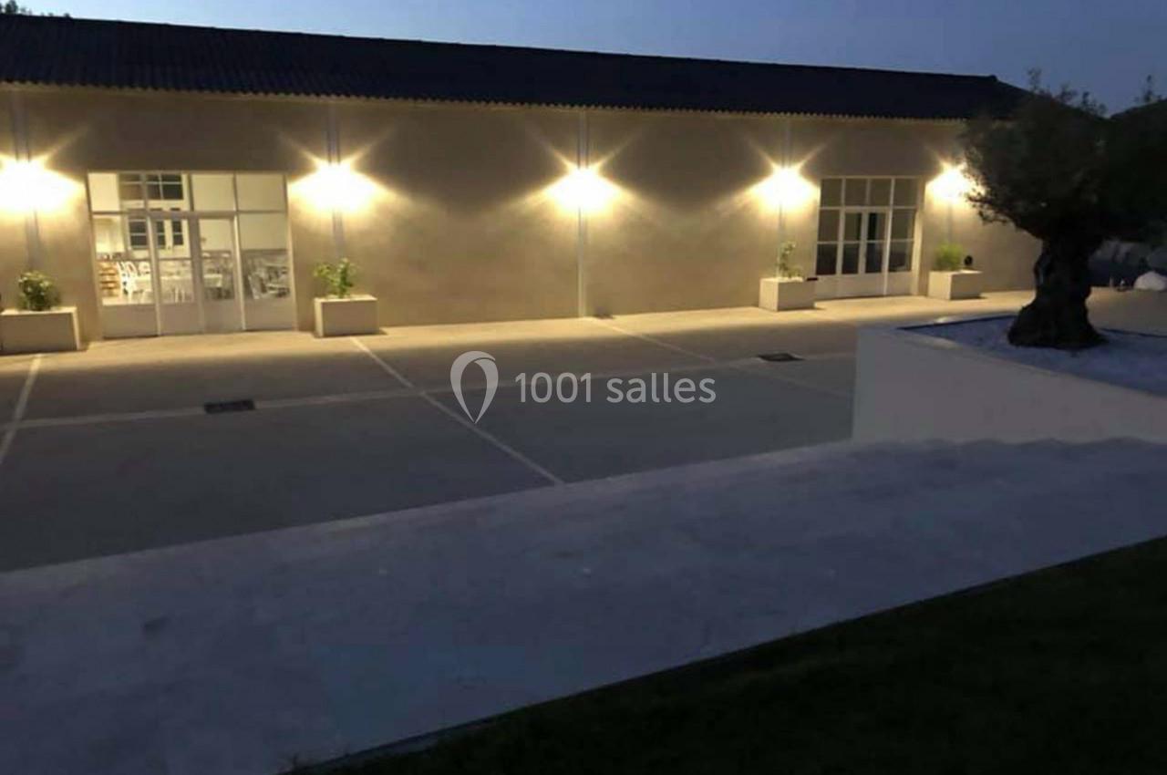 Façade d'un bâtiment éclairé par des appliques murales, avec une cour dégagée et un olivier en pot au premier plan.