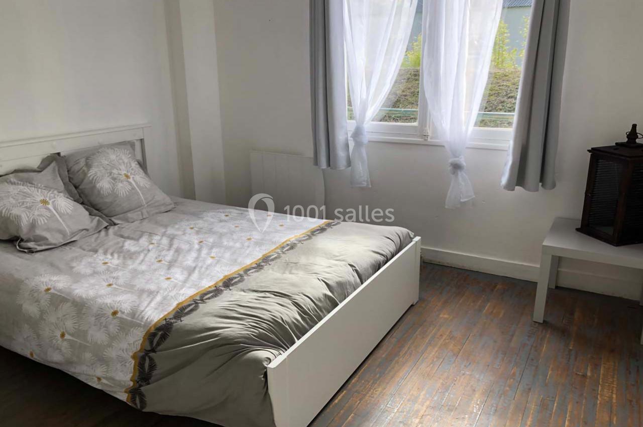 Chambre lumineuse avec lit double, draps à motifs, parquet en bois et fenêtre avec rideaux blancs.