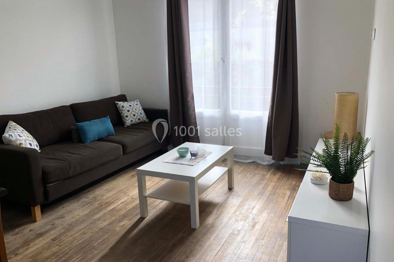 Salon lumineux avec canapé marron, table basse blanche, parquet en bois et fenêtre avec rideaux.