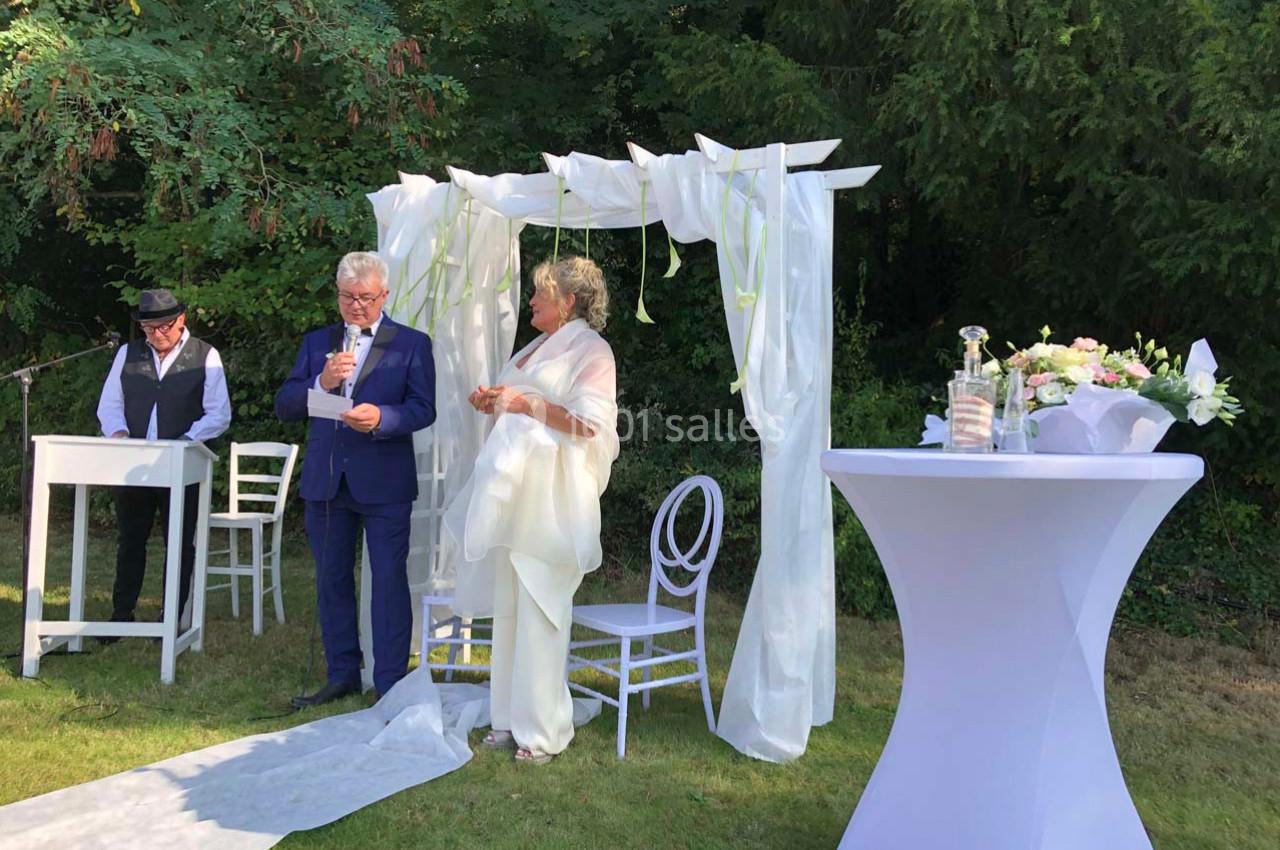 Un couple échange des vœux sous une arche blanche décorée, dans un jardin, avec un officiant à leurs côtés.