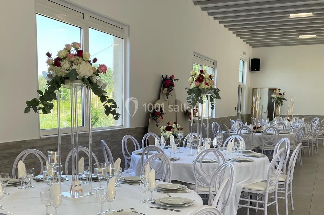Salle de réception lumineuse décorée avec des tables rondes, nappes blanches et centres de table fleuris.