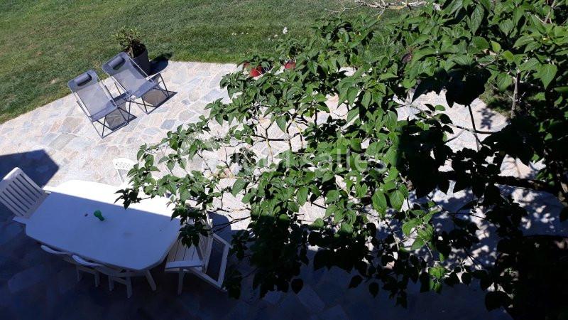 Terrasse en pierre avec table blanche, chaises et arbre ombrageant partiellement la scène, vue sur pelouse.