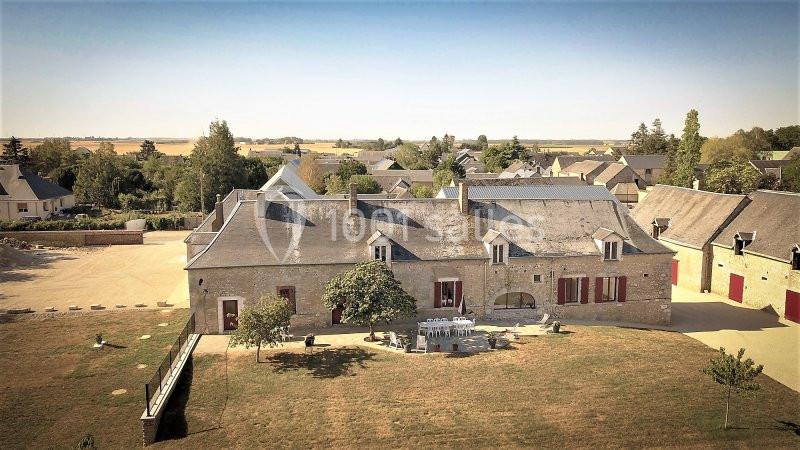 Bâtisse en pierre avec volets rouges, entourée de pelouse et d'arbres, dans un paysage rural ensoleillé.
