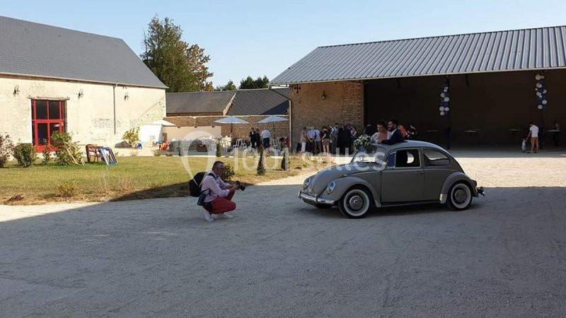 Une voiture ancienne grise décorée pour un événement est photographiée dans une cour avec des invités en arrière-plan.