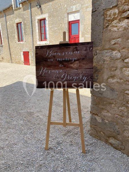 Panneau en bois sur chevalet indiquant ’Bienvenue au mariage de Roxane & Grégory’, placé dans une cour ensoleillée.